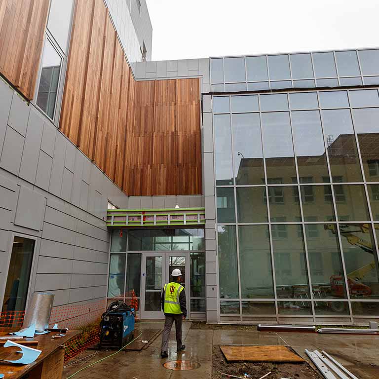 Indiana University construction worker stands near building materials and looks up at a large window. 